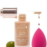 A bottle of 100% Pure 2nd Skin Foundation with a dropper dispensing the natural coverage serum sits on a white background beside a pink makeup sponge and a green olive.