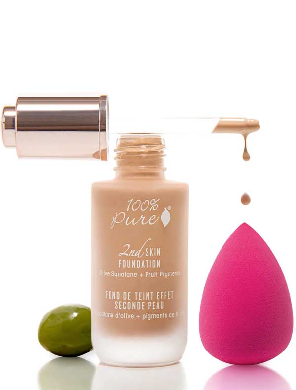 A bottle of 100% Pure 2nd Skin Foundation with a dropper dispensing the natural coverage serum sits on a white background beside a pink makeup sponge and a green olive.