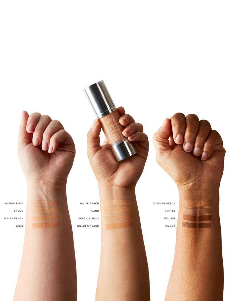 Three arms with different skin tones display swatches of 100% Pure Healthy Foundation in Alpine Rose, Crème, White Peach, Sand, Peach Bisque, Golden Peach, Toffee, Mousse, and Cocoa shades. The middle arm holds a foundation bottle.