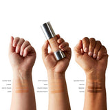 Three arms with different skin tones display swatches of 100% Pure Healthy Foundation in Alpine Rose, Crème, White Peach, Sand, Peach Bisque, Golden Peach, Toffee, Mousse, and Cocoa shades. The middle arm holds a foundation bottle.