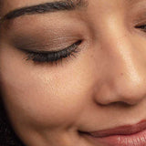 A close-up of a person’s face with closed eyes, showcasing shimmery brown tones from the 100% Pure Better Naked Palette 9g, black eyeliner, long lashes, defined brows, and a soft dimpled smile.