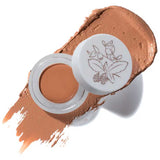 An open container of 100% Pure Long Last Concealer 3g in Toffee (medium brown), shown on a textured swatch. The lid displays a botanical illustration.