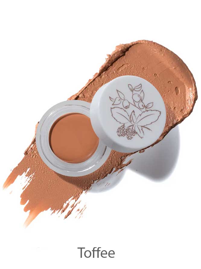 An open container of 100% Pure Long Last Concealer 3g in Toffee (medium brown), shown on a textured swatch. The lid displays a botanical illustration.